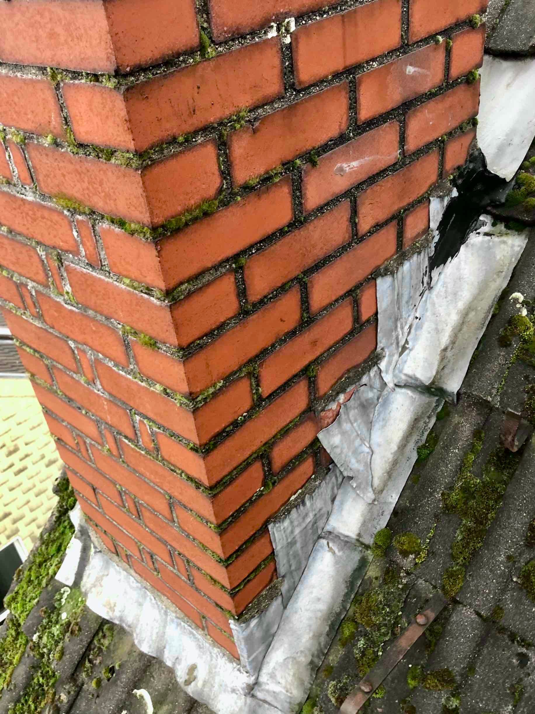A red brick chimney with moss in its mortar joints and flashing lifting at one corner